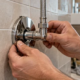 A plumber fits a chrome scutcheon plate around a water supply pipe through a tiled wall.