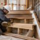 A carpenter replaces a timber stair tread while an inspector checks the tread support and handrail connection before the stair reopens.