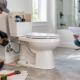 A plumber installs and checks a toilet connection while an inspector reviews the bathroom rough-in plan.