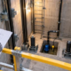 An engineer reviews an elevator pit drawing from behind guardrails while checking the sump pump, float switch, rails, and buffers below.
