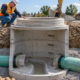 A precast sanitary sewer manhole connects two pipes in a shored trench as an engineer checks the rim.