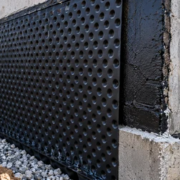 A worker fastens a dimpled drainage board over a waterproofed foundation above a gravel footing drain.