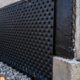 A worker fastens a dimpled drainage board over a waterproofed foundation above a gravel footing drain.