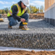 A construction engineer checks a compacted gravel subbase above geotextile and a perforated drain beside a concrete foundation.