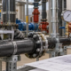 An inspector measures a commercial pipe with a caliper while a technician checks its pressure gauge against the drawing.