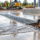 A construction worker lifts a trench drain grate to clear debris while runoff flows from a sloped paved jobsite.