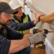 A carpenter tightens the wall-mounted handrail bracket while an inspector checks the rail for movement in a closed stair repair area.
