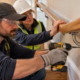 A carpenter tightens the wall-mounted handrail bracket while an inspector checks the rail for movement in a closed stair repair area.