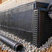 A worker installs dimpled drain board over waterproofing on a basement foundation wall above gravel and a footing drain.