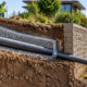 Perforated interceptor drain in gravel beside a retaining wall, routing hillside water to a safe outlet.