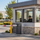 Security guard monitors a glass gatehouse and vehicle barrier at a commercial property entrance.