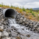 A drainage pipe daylight outlet releases water onto a rock-lined apron at a graded site.