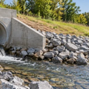 A civil engineer checks a concrete stormwater pipe outfall as water discharges into a riprap-lined channel that protects the receiving bank from erosion.
