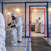 Remediation workers in protective suits remove material inside sealed plastic containment with HEPA negative-air equipment.