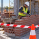 Bricklayer works inside a barricaded masonry limited-access zone on a construction site.