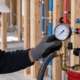 Plumber checking an analog pressure gauge on a test pump connected to red and blue water lines before walls close.