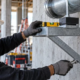 A construction worker levels a galvanized bracket anchored to a concrete wall to support mechanical equipment.