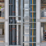 An inspector reviews plumbing, ductwork, and cable routes inside a building services shaft.