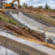 A site engineer traces runoff from a grated drain through a graded channel to a protected outlet at a construction site.