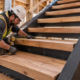 A carpenter checks the fit and depth of a broad wood stair tread on steel stringers while an engineer reviews the landing connection.