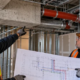 A site engineer and trade leads compare a pipe route blocked by a structural beam with their coordination drawing.