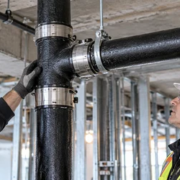An inspector checks the shielded coupling and hanger support on a commercial cast-iron drainage pipe installation.