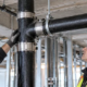 An inspector checks the shielded coupling and hanger support on a commercial cast-iron drainage pipe installation.