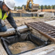 Construction workers inspect a geotextile silt sack inside a storm drain inlet to capture sediment from runoff.