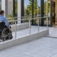 A wheelchair user approaches a public entrance along a guarded ramp with a level landing as engineers review the door transition.