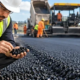 Road engineer inspects a dark asphalt sample beside a freshly paved roadway and roller.