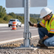 Road crew member inspects the breakaway coupling at the base of a roadside sign support.