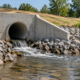 An engineer inspects a stormwater outfall headwall and riprap apron where a drainage pipe discharges.