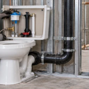 A plumber checks the toilet cistern flush valve and water supply while an inspector reviews the waste connection before close-in.