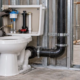 A plumber checks the toilet cistern flush valve and water supply while an inspector reviews the waste connection before close-in.