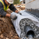 A worker wraps permeable geotextile around a gravel-filled perforated drainpipe beside a foundation, keeping surrounding soil out of the drainage stone.