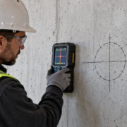 Construction worker checking a marked concrete wall opening with a handheld scanner beside an orange-sleeved service penetration.