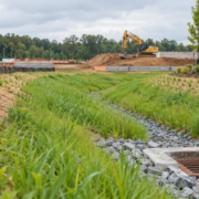 A site engineer checks silt fencing and a vegetated swale carrying runoff toward a protected catch basin to reduce construction sediment.