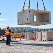 A crane crew positions a precast concrete element with checked slings, a tagline, and a signaler outside the load path.