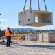 A crane crew positions a precast concrete element with checked slings, a tagline, and a signaler outside the load path.