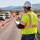 A work-zone safety supervisor checks traffic signs, cones, and barriers while vehicles pass a protected construction area.