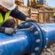 A construction engineer checks a bolted flange joint connecting blue water pipes in a shored trench with a torque wrench.