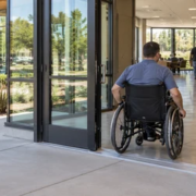 A wheelchair user enters a public building along a clear accessible route as an accessibility consultant reviews the entrance and interior circulation.
