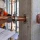 Plumbing and fire-stop crews coordinate a sleeved pipe passing through a concrete wall while checking drawings and seal clearance.