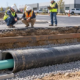 A drainage pipe runs through a larger protective casing beneath a commercial access road as an engineer checks alignment before backfill.