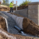 A site engineer and worker inspect a gravel interceptor drain beside a retaining wall, with a perforated pipe directing runoff away from the structure.