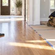 A worker vacuums a hardwood floor while another lays taped protective sheets to guard the finish from grit and construction traffic.