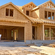 Builders reviewing house plans beside a framed two-story home on a residential construction site.
