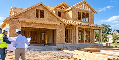 Builders reviewing house plans beside a framed two-story home on a residential construction site.