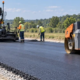 A paving crew lays fresh asphalt while a road roller compacts the new surface along the roadway.