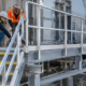 Installers inspect the guarded steel catwalk stair entry and gate while checking support brackets and the route onto the platform.